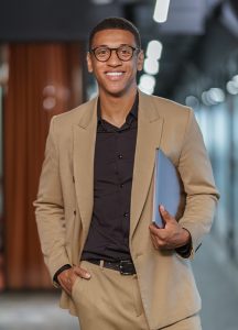Hombre joven con traje beige y camisa negra, sonriente, sostiene una carpeta en la mano, en un entorno de oficina moderna con iluminación brillante y paredes de vidrio.