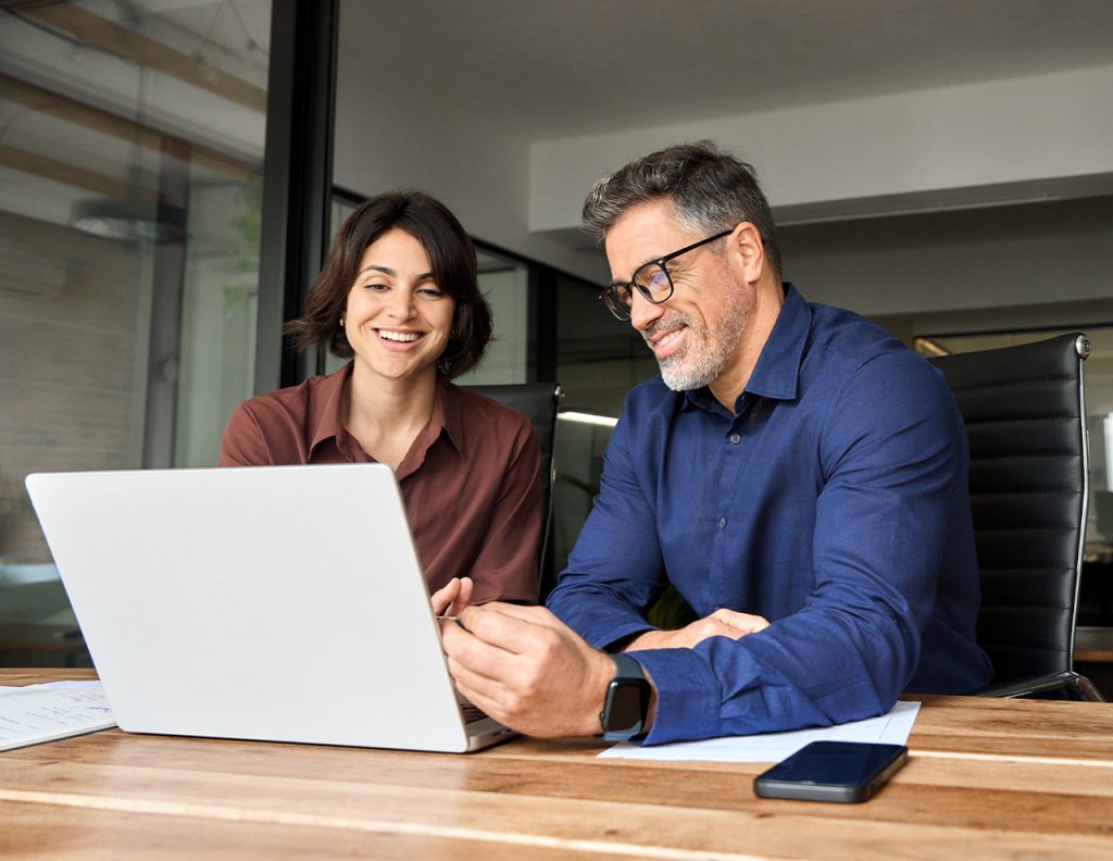 Hombre y mujer sonríen mientras trabajan juntos en una computadora portátil en una mesa de madera. El entorno es moderno y luminoso, con sillas negras y documentos en la superficie.
