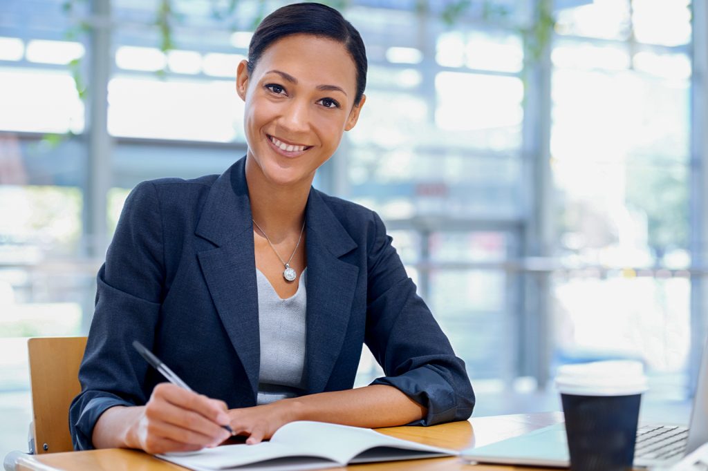 Mujer sonriente con traje oscuro, escribiendo en un cuaderno sobre una mesa. Fondo luminoso con plantas y un ordenador portátil junto a una taza de café.