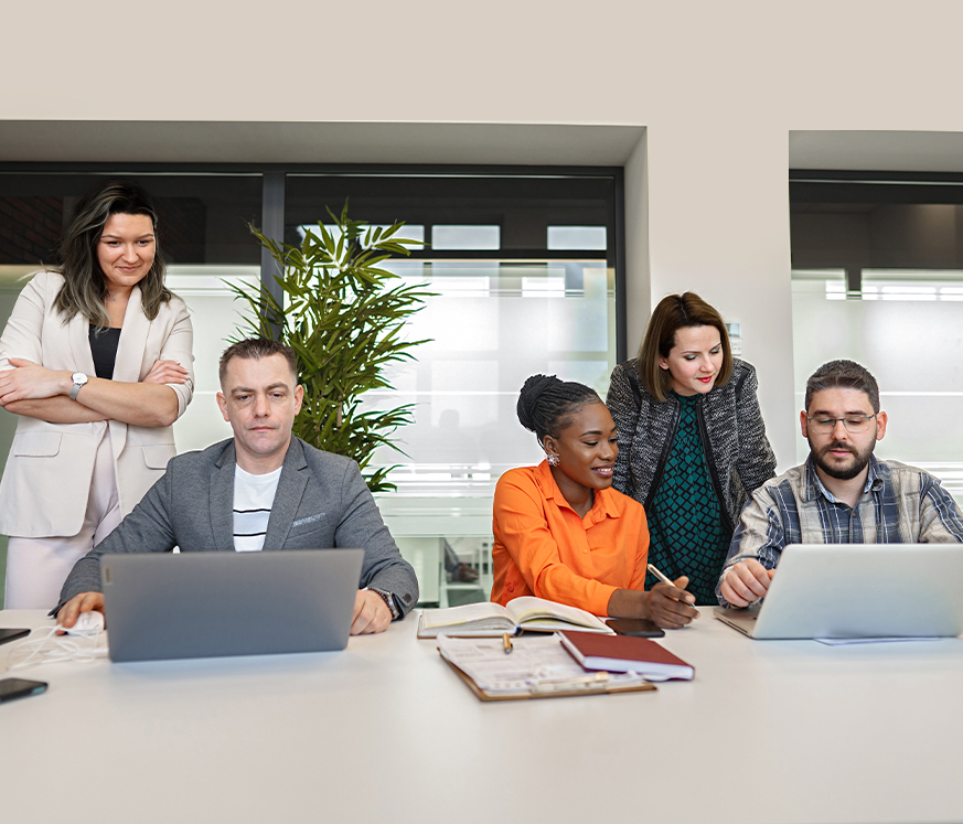 Grupo de cuatro personas sentadas alrededor de una mesa, trabajando en laptops y tomando notas. Hay una planta al fondo y un entorno luminoso. Predominan colores neutros y naranjas.