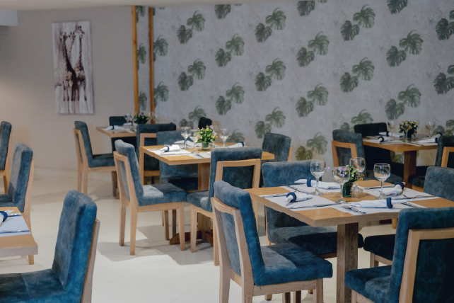 Restaurante con mesas de madera y sillas de terciopelo azul, decorado con papel tapiz de hojas verdes. Flores y copas de cristal en las mesas, iluminación suave.