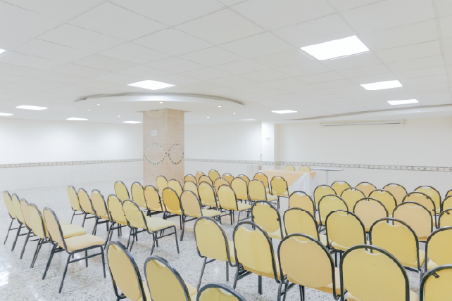 Salón con filas de sillas amarillas dispuestas en un espacio iluminado, con paredes blancas y un pilar central decorado. Entorno limpio y ordenado, sin personas visibles.