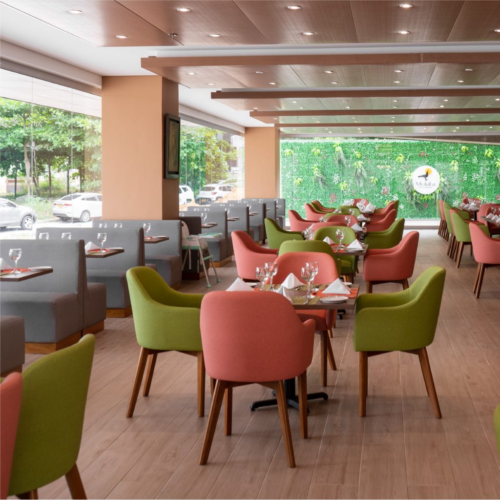 Restaurante con mesas y sillas en colores verde y rosa, decorado con plantas en la pared y ventanales que permiten la entrada de luz natural.