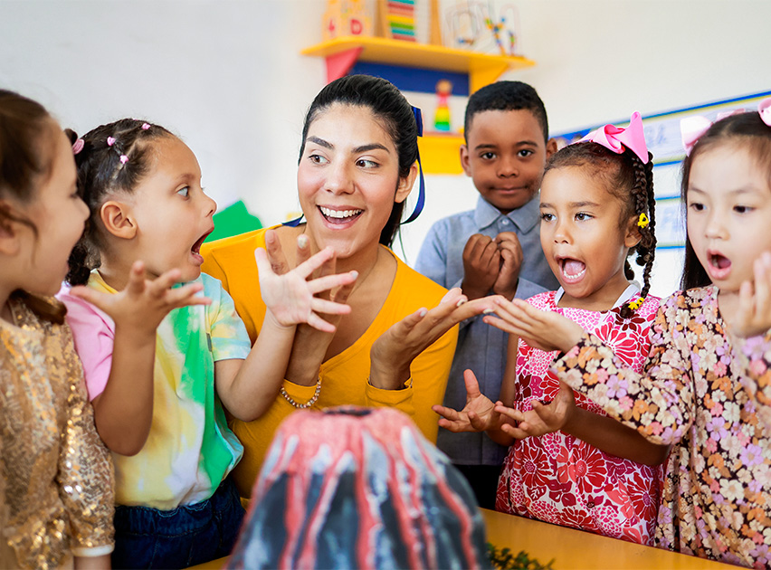 Grupo de niños y una mujer sonriente interactuando alrededor de un volcán de juguete en un aula colorida, mostrando expresiones de sorpresa y entusiasmo.