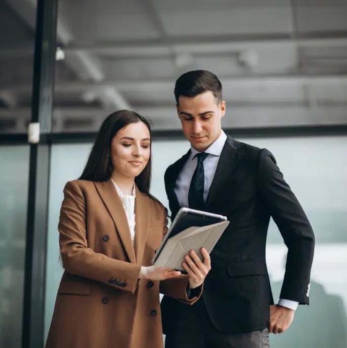Dos personas en un entorno de oficina moderno, revisando una tablet. La mujer lleva un abrigo marrón y el hombre un traje negro. Predominan tonos neutros en el diseño.