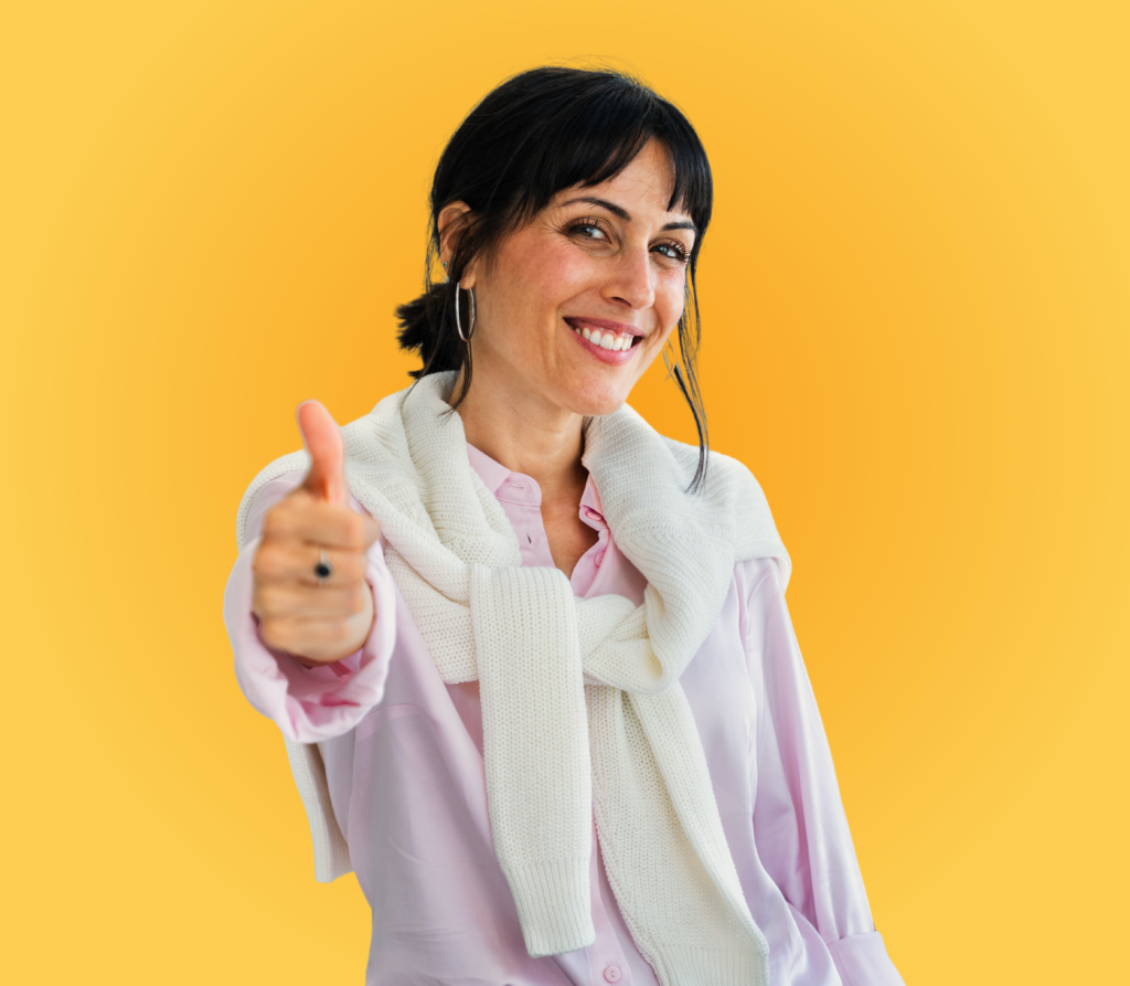 Mujer sonriente con blusa rosa y suéter blanco atado al cuello, levantando el pulgar en señal de aprobación, con fondo amarillo.