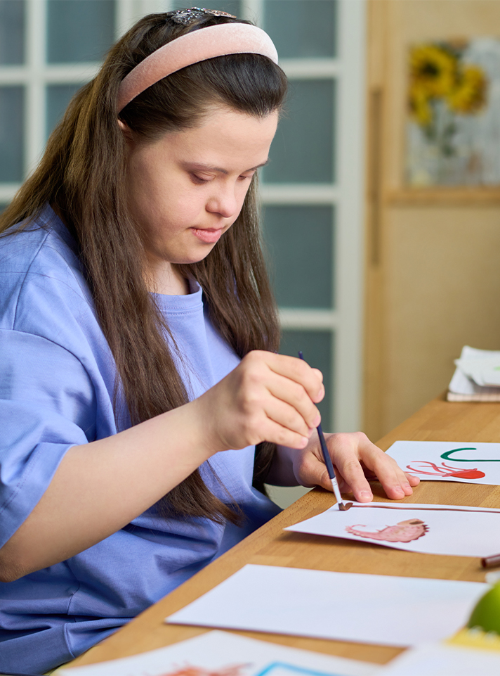 Joven con cabello largo y castaño, usando una camiseta azul, pinta en una mesa de madera. Hay varios dibujos en papel y un ambiente iluminado con flores al fondo.
