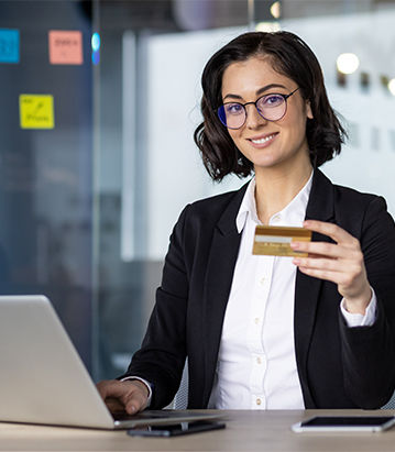 Mujer con gafas sonriente sosteniendo una tarjeta de crédito, sentada frente a una laptop en un entorno de oficina moderna, con post-its de colores en el fondo.