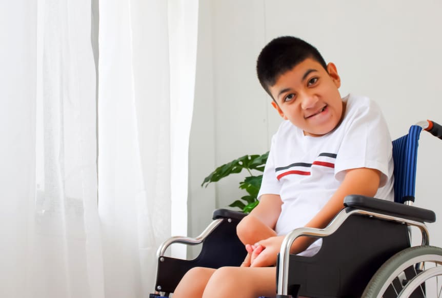 Niño en silla de ruedas, sonriendo, con camiseta blanca y rayas, junto a una planta y cortinas blancas.