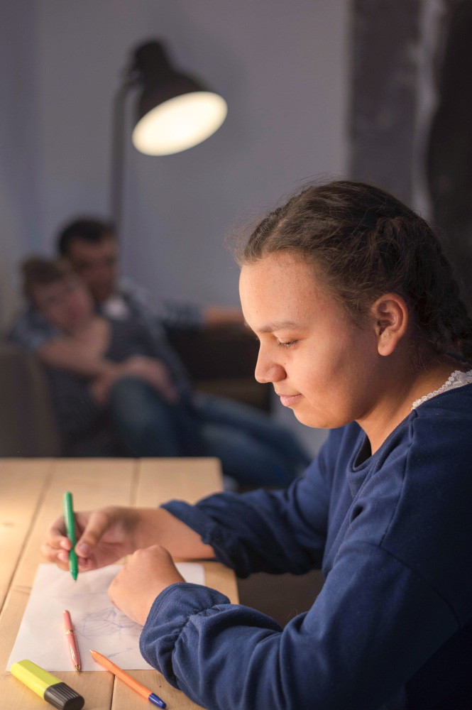 Niña dibujando en una mesa con lápices de colores, iluminada por una lámpara. Al fondo, dos personas sentadas en un sofá. Colores suaves y ambiente acogedor.