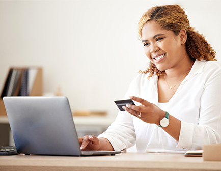 Mujer con cabello rizado y suelto sonríe mientras sostiene una tarjeta de crédito y usa una laptop. El entorno es luminoso y moderno, con muebles de madera clara.